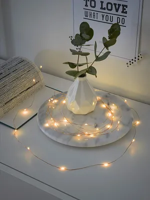 A string of copper fairy lights is draped around a white vase with green foliage on a marble tray, placed on a white surface.