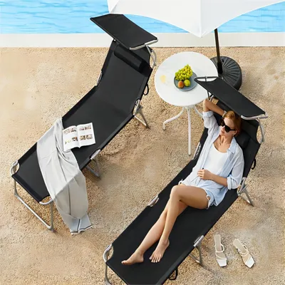 Two black lounge chairs are set up on a sandy area next to a swimming pool. One chair has a book and a blanket on it, while the other has a woman relaxing on it. A small white table with fruit and a white umbrella are also present.