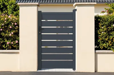 A modern gray aluminum garden gate with horizontal slats, set between two cream-colored pillars and surrounded by greenery.