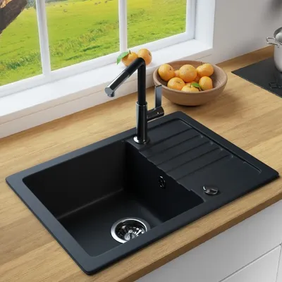 A black kitchen sink with a faucet and a bowl of oranges on a wooden countertop, with a window in the background showing a green field.