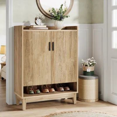 A wooden shoe cabinet with doors and an open shelf for shoes, placed in a hallway next to a round mirror and a small stool with flowers.