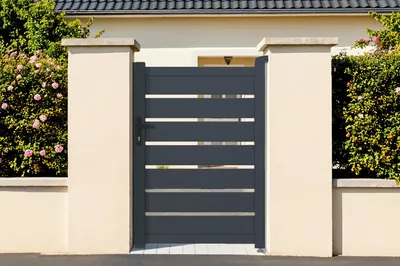 A modern gray aluminum gate and matching garden gate are shown in front of a house, flanked by beige pillars and lush green bushes.