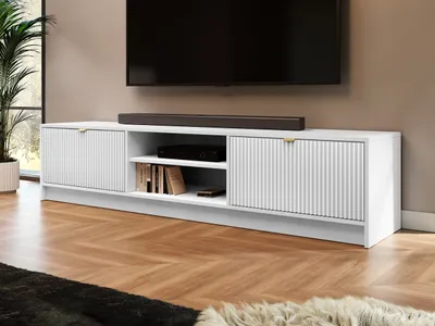 A white TV stand with fluted doors and gold handles, placed on a wooden floor in front of a wall. A large black TV is mounted on the wall above the stand.
