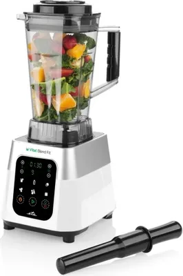 A white blender with a transparent jug filled with fruits and vegetables, placed on a white background. A black accessory is placed next to it.