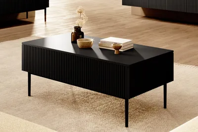 A black coffee table with fluted sides and thin metal legs is placed on a textured rug in a living room setting. Decorative items like a vase with dried flowers, a bowl, and books are placed on top of the table.
