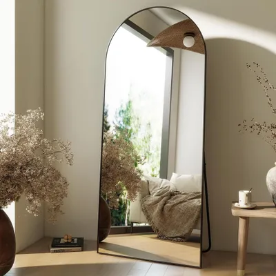 A full-length arched mirror is placed in a room with natural light coming from a window. Decorative dried flowers and a small table with a cup are also visible.