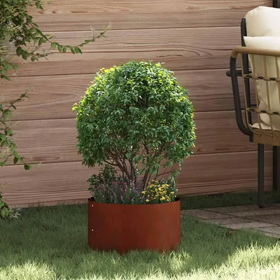 A round, rusty metal planter with a green bush and smaller purple and yellow flowers, placed on grass next to a wooden fence and a patio.