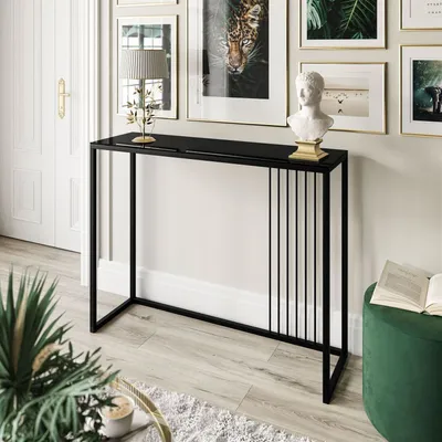 A modern black console table with a glass top, set against a neutral wall with framed artwork and a white door. Decorative items like a bust and a table lamp are placed on the table.
