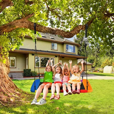 Four young children are happily sitting on a colorful rectangular swing attached to a large tree branch in a backyard, with a house in the background.
