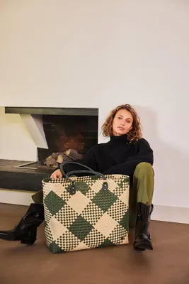 A woman sits on the floor next to a fireplace, holding a large checkered tote bag.