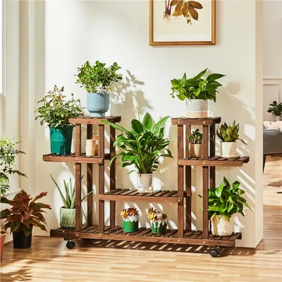 A multi-tiered wooden plant stand filled with various potted plants, placed indoors against a white wall.