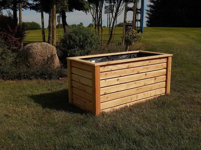 A wooden planter box sits on a grassy lawn in a garden setting, with trees and bushes in the background.