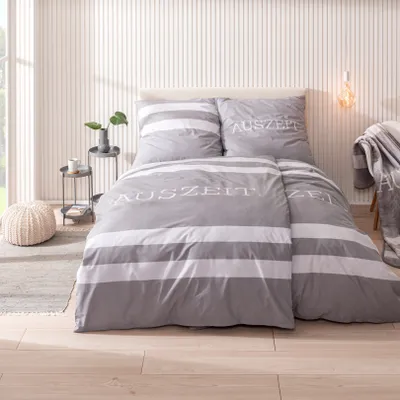 A neatly made bed with grey and white striped bedding, featuring the word "Auszeit" embroidered on the duvet and pillow.