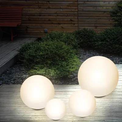 Four illuminated spherical garden lights of varying sizes are placed on a wooden deck in front of a wooden fence and green bushes.