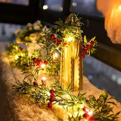 A festive garland with red berries and green leaves, illuminated by warm LED lights, is draped along a fuzzy surface, possibly a windowsill or mantelpiece, with a blurred background of lights.