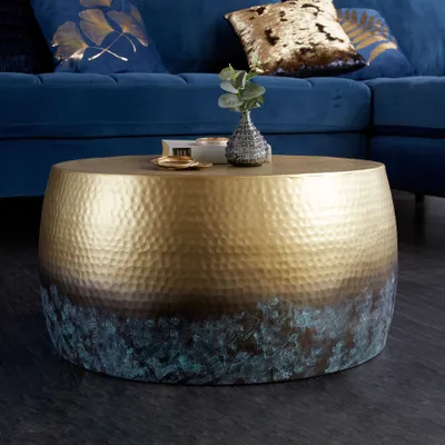 A round coffee table with a hammered gold and patina finish is placed in a living room setting, next to a blue sofa with decorative pillows.