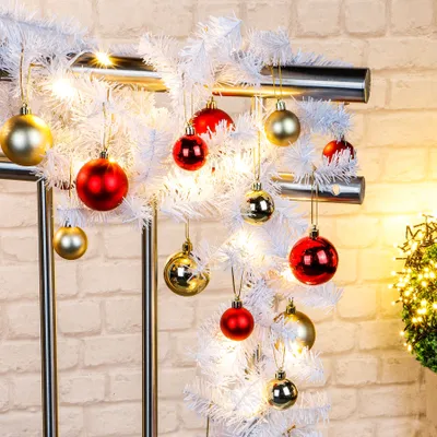 A white Christmas garland decorated with red and gold baubles and warm white fairy lights, set against a brick wall.