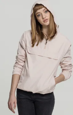 A young woman wearing a light pink hooded windbreaker and dark jeans, posing against a plain light gray background.