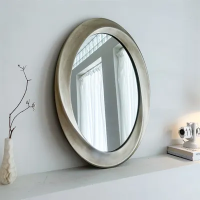 An oval mirror with a decorative frame is displayed on a shelf, reflecting a window with white curtains.