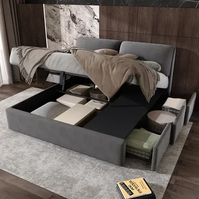 A modern bedroom with a grey upholstered bed frame, featuring a lifted mattress to reveal storage space underneath. The bed is dressed with beige and grey blankets and pillows. The headboard is against a wall with marble and wood paneling.