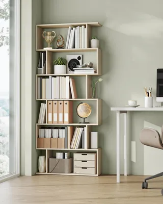 A modern, light wood bookshelf filled with books and decorative items, placed next to a window and a desk.