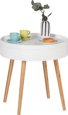 A round white side table with three wooden legs, featuring two cups of coffee, a pastry, and a vase with greenery.