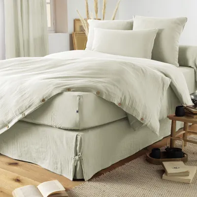 A bed with a light green duvet and bed skirt, styled in a bedroom setting with curtains and decorative items.