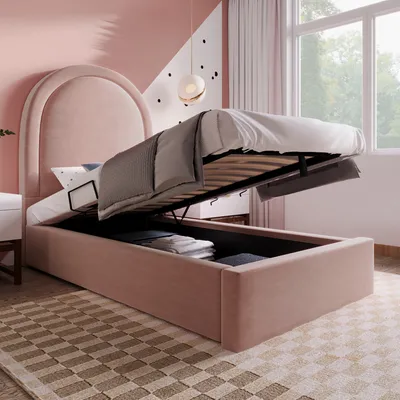 A bedroom scene with a pink upholstered bed frame that has its storage compartment open, revealing folded linens inside. The bed is made with gray and white bedding.