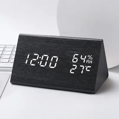 A black wooden digital alarm clock displaying the time, humidity, and temperature is placed on a white desk next to a keyboard.