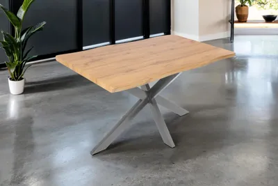 A wooden dining table with a metal spider base is shown in a room with a concrete floor and a dark wall.