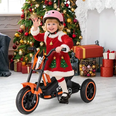 A young child dressed in a Santa-themed outfit rides an orange electric tricycle in front of a decorated Christmas tree and presents.