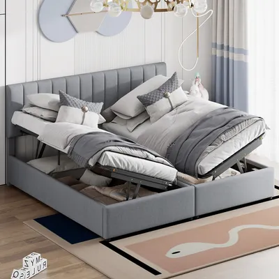 A bedroom scene with a grey upholstered bed frame, featuring two open storage compartments revealing bedding and storage items. The bed is made with white and grey linens and pillows. A decorative wall art and curtains are visible in the background.