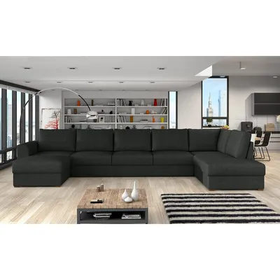A large, dark gray sectional sofa is shown in a modern living room setting with a bookshelf and large windows in the background.