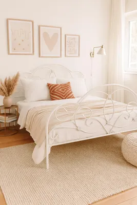 A bedroom scene with a white metal bed frame, white bedding, and decorative pillows and throws. Artwork is displayed on the wall above the bed, and a rug covers the floor.