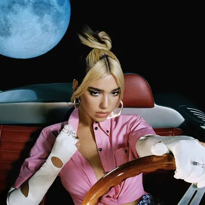 A woman in a pink shirt and white gloves sits in a car with a large blue moon in the background.