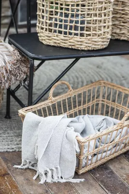 A woven basket filled with a folded blanket sits on a wooden floor, with another woven basket on a black bench in the background.