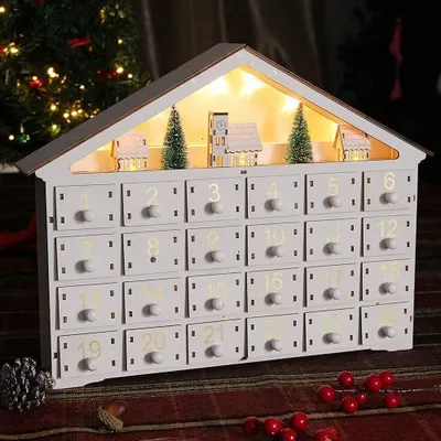 A white wooden advent calendar shaped like a house, decorated with miniature Christmas trees and houses, illuminated by fairy lights, set against a festive background with a Christmas tree and red berries.