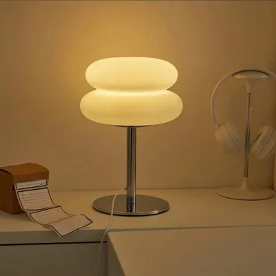 A table lamp with a dome-shaped shade sits on a white surface, next to a box with papers and a pair of headphones on a stand.