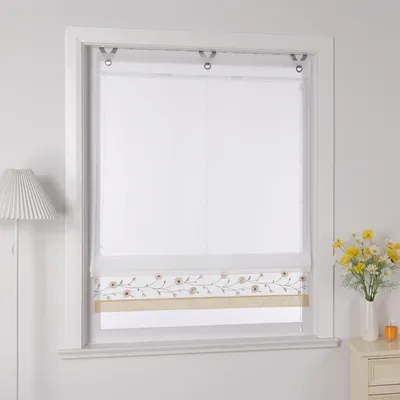 A white Roman blind with a floral embroidery detail is shown hanging in a window frame. A lamp is visible to the left, and a vase of yellow flowers sits on a nightstand to the right.