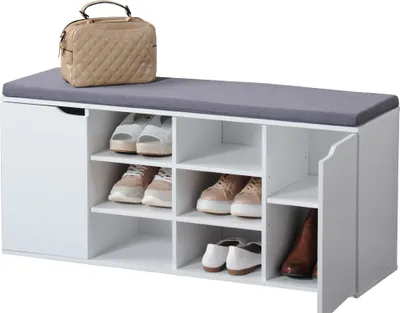 A white shoe storage bench with a gray cushioned seat, a beige handbag on top, and various shoes stored inside.