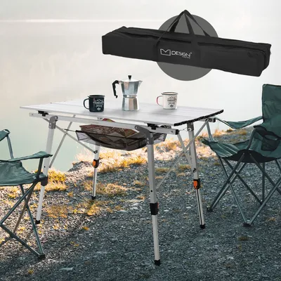 A camping table and chairs set up outdoors by a body of water, with a carrying bag shown above.