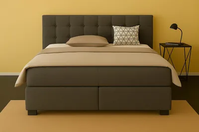 A boxspring bed with a dark gray upholstered headboard and base, set against a yellow wall with a small black side table and lamp.