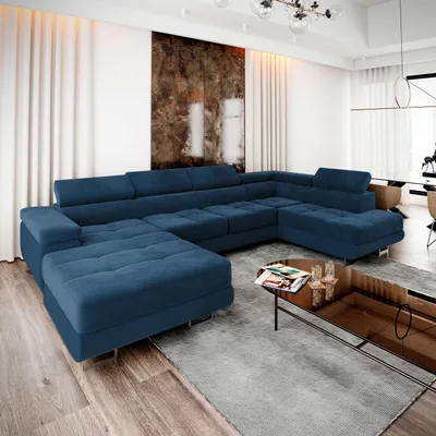A large, sectional blue sofa is arranged in a modern living room with sheer white curtains in the background and a large abstract artwork on the wall. A coffee table with magazines and glasses sits in front of the sofa.
