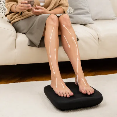A person sitting on a couch with their feet on a black foot massager, with arrows indicating massage flow on their legs.
