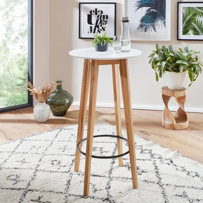 A round white bar table with wooden legs is shown in a room with a rug and decorative items.