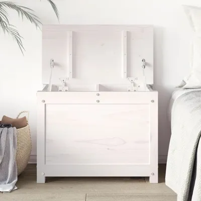 A white wooden storage chest with its lid open, placed next to a bed and a basket of laundry.