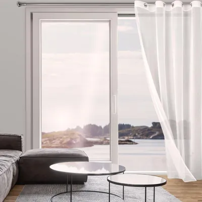 A living room with a large sliding glass door overlooking a body of water and islands. A white sheer curtain is partially drawn.