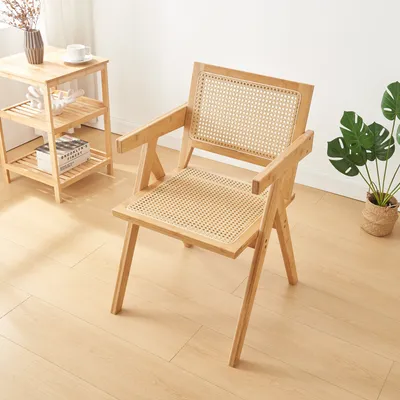 A wooden chair with rattan seat and backrest is placed next to a small wooden side table with a vase and a cup on it. A potted plant is visible in the background.