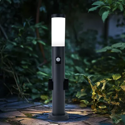 A dark gray bollard light with a motion sensor is shown in a garden setting at dusk, with plants and a stone path visible.