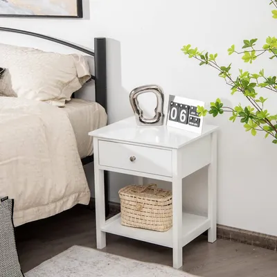 A white nightstand with a drawer and an open shelf is placed next to a bed.